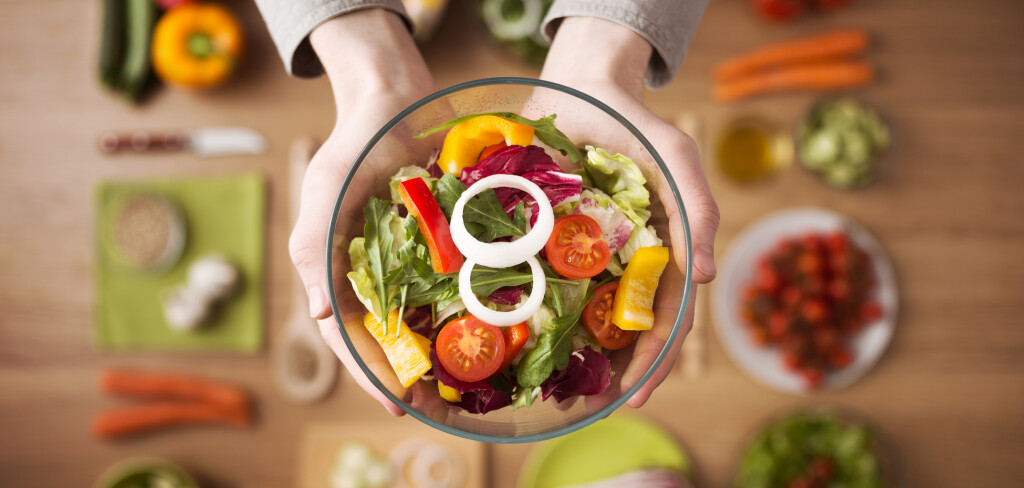 Hands holding an healthy fresh vegetarian salad in a bowl, fresh raw vegetables on background, top view