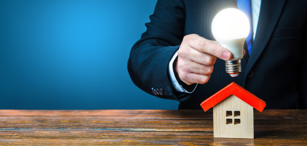Man is holding a glowing light bulb over the house. Smart home concept. Provision of housing with electricity and communications. Deal contract with supply utilities companies. Pay electricity bills