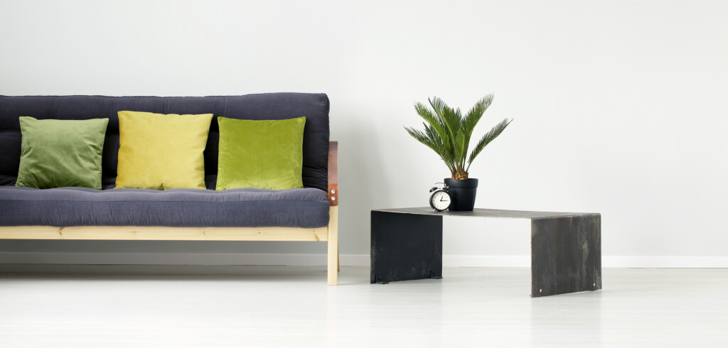 Plant and clock on metal table next to a black settee with green and yellow pillow in flat interior with empty wall