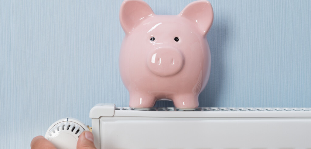 Close-up Of Man's Hand Adjusting Thermostat With Piggy Bank On Radiator