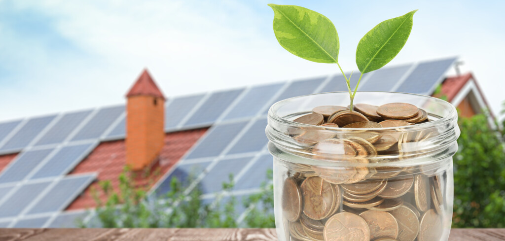 Glass jar with coins and plant against house with installed solar panels on roof. Economic benefits of renewable energy