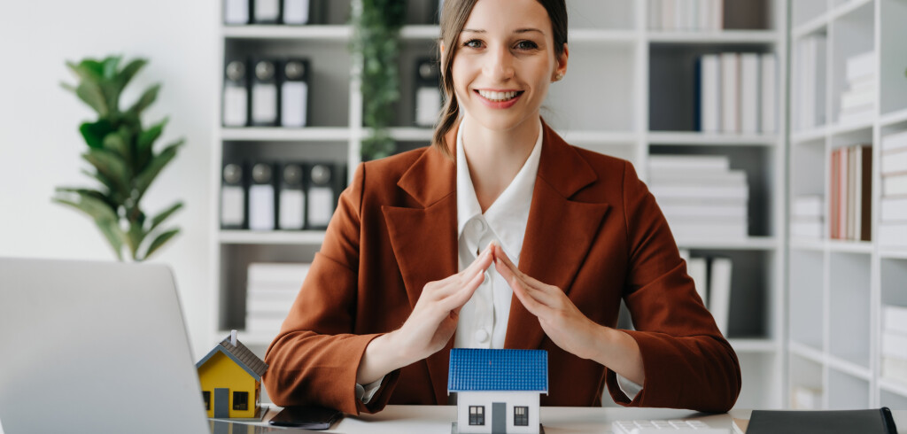 Caucasian Female real estate agent is use hands to protect red roof for the concept of real estate investment about house trading, purchase at desk in office