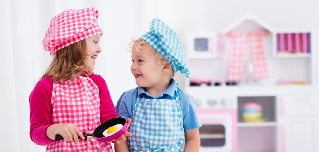 Little girl and boy in chef hat and apron cooking fried eggs in toy kitchen.  Wooden toys for young children. Kids play and cook at home or daycare. Toddler kid playing with stove, pans and dishes.